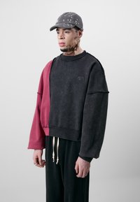 Schwarzes und burgunderfarbenes übergroßes Sweatshirt mit einem rohen Kanten-Design an den Ärmeln, ausgestattet mit einem Logo und cremefarbenen Kordeln. Kombiniert mit schwarzen Hosen.