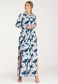 Długa, kwiecista sukienka maxi z długimi rękawami, kołnierzykowym dekoltem i wysokimi rozcięciami. Posiada granatową bazę z białymi i zielonymi wzorami kwiatowymi.