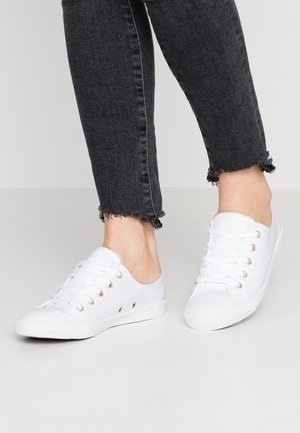 Baskets basses en toile blanche avec lacets blancs portées avec un jean skinny noir à ourlet effiloché sur un fond neutre.