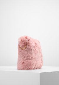 Růžová kabelka z umělé kožešiny zaobleného tvaru s pozlaceným kroužkem na boku. Měkká textura a hebký vzhled.