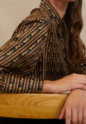 Femme portant un chemisier noir à rayures florales verticales, assise avec les bras reposant sur une surface en bois clair, longs cheveux bruns visibles.
