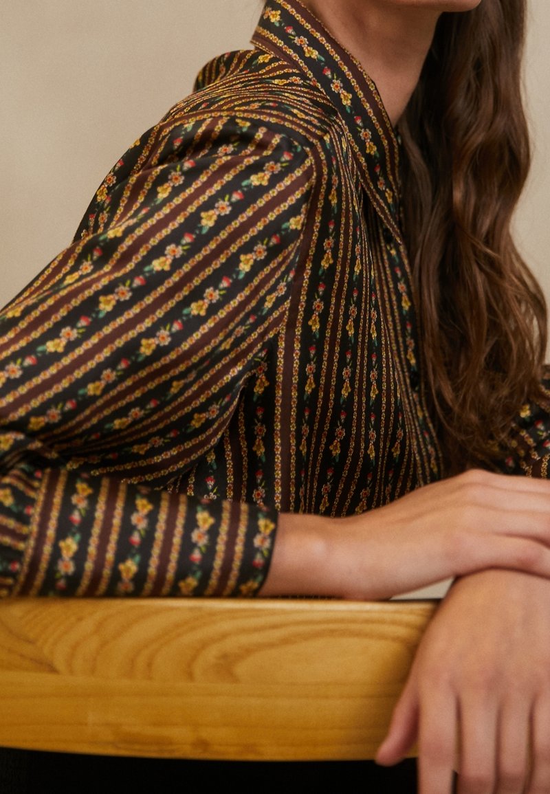 Femme portant un chemisier noir à rayures florales verticales, assise avec les bras reposant sur une surface en bois clair, longs cheveux bruns visibles.