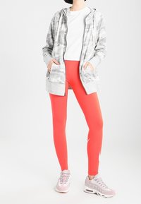 Osoba v bílé tričku, šedé maskáčové zipové bundě, jasně červených legínách a světle růžových teniskách stojí s rukama v kapsách.