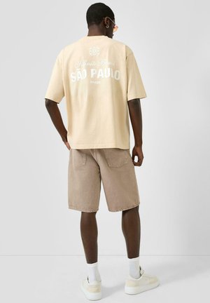 Mann trägt beige T-Shirt mit Aufdruck "São Paulo Brazil", hellbraune Jeansshorts, weiße Socken und cremeweiße Sneakers, steht vor einem einfarbigen Hintergrund.