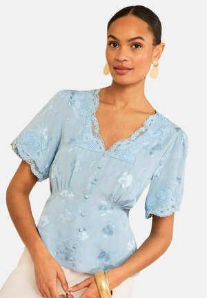 Kvinde iført en lyseblå blomstret bluse med blonder, pufærmer og stofbetrukne knapper, kombineret med beige bukser og gyldne accessories.