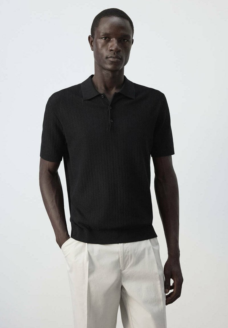 Homme portant un polo en maille noire à manches courtes et un pantalon beige clair, debout devant un fond clair uni.