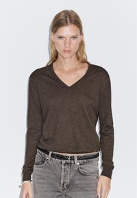 Brown long-sleeve V-neck top in a soft, textured material, paired with faded gray jeans and a braided black belt.