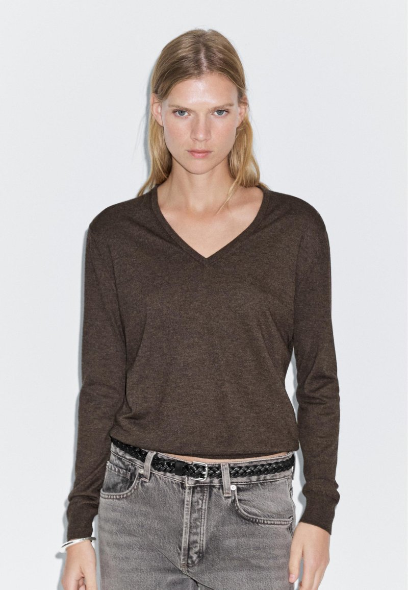 Brown long-sleeve V-neck top in a soft, textured material, paired with faded gray jeans and a braided black belt.