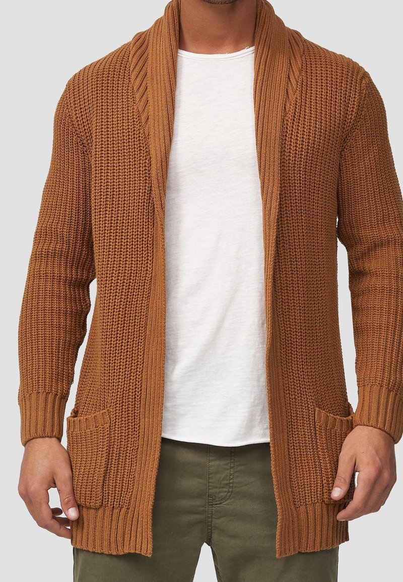 Gebreide bruine cardigan met een sjaalkraag, ribgebreide manchetten en twee fronttassen, met een textuurpatroon en een open voorkant.