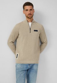 Beige Strickpullover mit hohem Kragen und kurzer Reißverschluss. Verfügt über eine dunkelblaue Akzenttasche auf der Brust und ein Seitenpatch-Detail.
