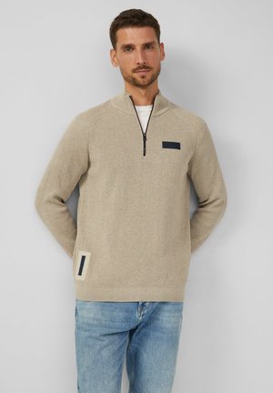 Beige Strickpullover mit hohem Kragen und kurzer Reißverschluss. Verfügt über eine dunkelblaue Akzenttasche auf der Brust und ein Seitenpatch-Detail.
