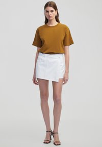 Bruine T-shirt met korte mouwen, gecombineerd met een witte denim minirok met voorzakken en een zijsluiting met knopen. Zwarte bandjesandalen zijn gedragen.