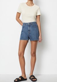 T-shirt a coste beige chiaro abbinato a pantaloni shorts in denim blu a vita alta. Sandali neri con dettagli in fibbia. Outfit semplice e casual.