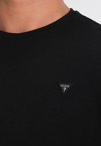 T-shirt en coton noir avec un col rond et un petit patch logo triangulaire portant le mot "GUESS" en blanc sur le côté gauche.
