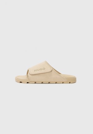 Beige sklidesandal med en myk tekstiloverside, borrelåsrem og teksturert gummisåle med rektangulært design og subtil logodetalj.