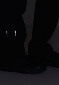 Jambes portant un pantalon noir et des chaussures de plein air noires avec des points réfléchissants blancs sur le pantalon près des mollets sur un fond sombre.