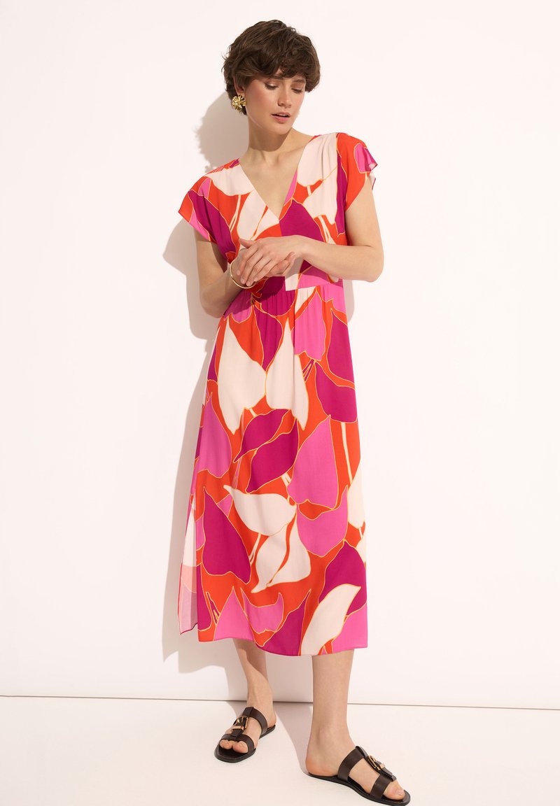 Kolorowa sukienka bez rękawów z kwiatowym wzorem w odcieniach różu, pomarańczy i bieli. O średniej długości z dekoltem w serek, zestawiona z czarnymi sandałami.