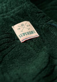 Tela de forro verde con superficie texturizada. Incluye una etiqueta crema con "SUPERDRY" en verde oscuro y un gráfico de ejes cruzados.