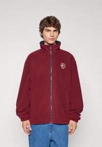 Giacca in pile borgogna con colletto alto, chiusura con zip e fodera blu. Presenta una toppa con logo e tasche laterali. Design dal taglio ampio.