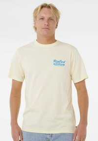Jonge man met blond haar, in een crèmekleurig Rip Curl Board Bakery t-shirt en lichtblauwe spijkerbroek, staande tegen een witte achtergrond.
