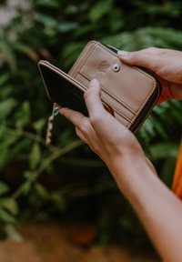 Compact wallet made of tan leather with zip closure, interior card slots, and a keychain; held by a hand against green foliage.
