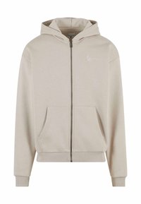 Beige zip-up hettegenser med frontlomme, ribbede mansjetter og kant, samt en liten hvit logo på brystet. Laget av mykt, teksturert stoff.