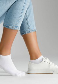 Weißes Canvas-Sneaker mit einer strukturierten Gummisohle neben einer weißen Knöchelsocke, getragen zu hellblauen, taillierten Jeans.