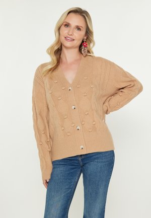 Tan gebreide cardigan met een V-hals, voorzien van gestructureerde patronen en grote knopen. Gecombineerd met blauwe jeans, tegen een effen achtergrond.