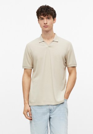 Junger Mann mit lockigem Haar, der ein beiges kurzärmeliges Poloshirt und hellblaue Jeans trägt, steht mit einer Hand in der Tasche.
