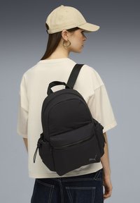 Sac à dos en tissu noir de forme arrondie, avec compartiments zippés et bretelles réglables. Porté avec un t-shirt de couleur claire, avec un logo latéral.