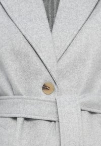 Cappotto di lana grigio chiaro con rever ampi, un grande bottone beige singolo, cintura in tessuto e zip visibile sotto.