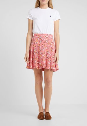 Vrouw draagt een wit T-shirt met korte mouwen en een roze bloemenrok gecombineerd met bruine instapschoenen, staand tegen een effen achtergrond.