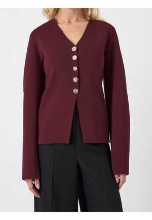 Femme portant un haut bordeaux à manches longues avec cinq boutons décoratifs dorés, associé à un pantalon noir à jambes larges.