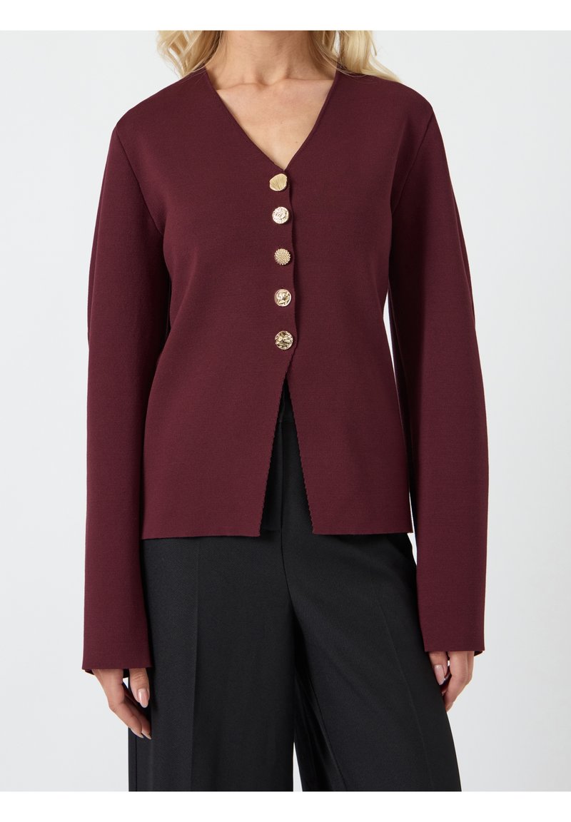 Femme portant un haut bordeaux à manches longues avec cinq boutons décoratifs dorés, associé à un pantalon noir à jambes larges.