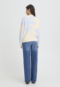 Beige neulepaita, jossa suuri vaaleansininen lehtikuvio, yhdistetty korkeavyötäröisiin sinisiin farkkuihin. Pehmeä materiaali, rento istuvuus ja kevyesti resoritut hihansuut.