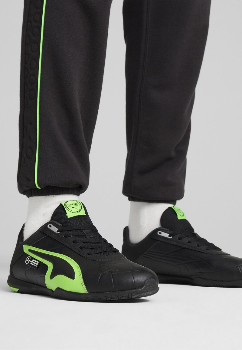 Chaussures de sport noires avec des accents verts, présentant une tige en cuir lisse, une semelle en caoutchouc texturée et une fermeture à lacets. Détails de logo remarquables inclus.