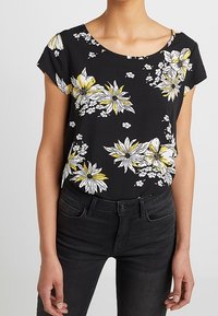 Blomstret sort top med korte ærmer; har gule og hvide blomster, en rund halsudskæring og et blødt, letstof. Sammen med sorte jeans.
