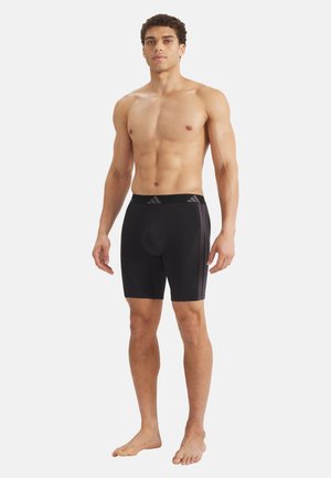 Homme musclé debout pieds nus portant un short de compression athlétique noir avec des bandes latérales et un logo à la taille sur fond uni.