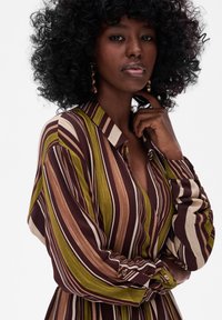 Koszula z długim rękawem i kołnierzykiem, z pionowymi paskami w kolorach burgundowym, zielonym i beżowym. Miękka tkanina z zapięciem na guziki przy mankietach i dekoltem w kształcie litery V.