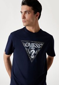 Navyblaues T-Shirt aus Baumwolle, mit einem dreieckigen Guess-Logo in der Mitte sowie Text und abgenutzten Designelementen. Kurze Ärmel.