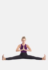 Top sportivo viola, leggings neri, eseguendo una posizione di split seduto su uno sfondo neutro, mani in posizione di preghiera, espressione concentrata.