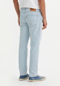 Man in lichtblauwe denim jeans en blauwe hoge sneakers, staande op een witte achtergrond met zijn rug naar de camera gekeerd.