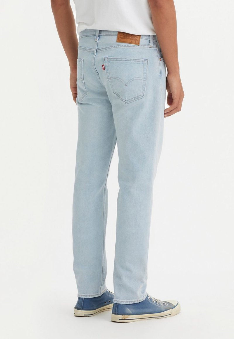 Homme portant un jean en denim bleu clair et des baskets montantes bleues, se tenant sur un fond blanc de dos.