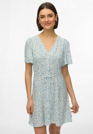 Kvinde iført en kort, hvid blomstret kjole med blå blomster, V-hals, korte ærmer og bindebånd i taljen, smilende med kort mørkt hår.