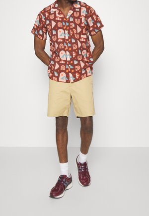 Camicia a maniche corte con sfondo marrone, caratterizzata da motivi geometrici e nuvole colorati; abbinata a pantaloni corti beige e sneakers bordeaux.