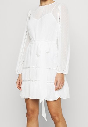 Hvid kjole med gennemsigtige lange ærmer, polkaprikket tekstur, bælte i taljen, lagdelte flæser og blondeaccenter på ærmer og krop.