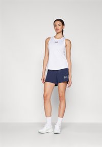 Top atlético branco sem mangas com logotipo, combinado com calções azul-marinho com logotipo, complementado por sapatos e meias atléticas brancas.
