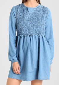 Denimmekko, jossa on rypytetty yläosa, pitkät hihat ja rypytetty helma. Vaalea sininen väri ja elastiset hihansuut tarjoavat rennon istuvuuden.