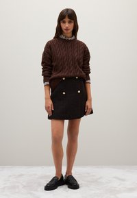 Bruine kabelgebreide trui over een zwarte tweed mini-jurk met gouden knopen. Zwarte instap schoenen, geruite kraag zichtbaar eronder.