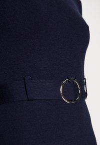 Mörkblått stickat tyg med en smidig, figurnära design som har ett bälte med en silverfärgad cirkulär spänne, vilket ger en dekorativ detalj.